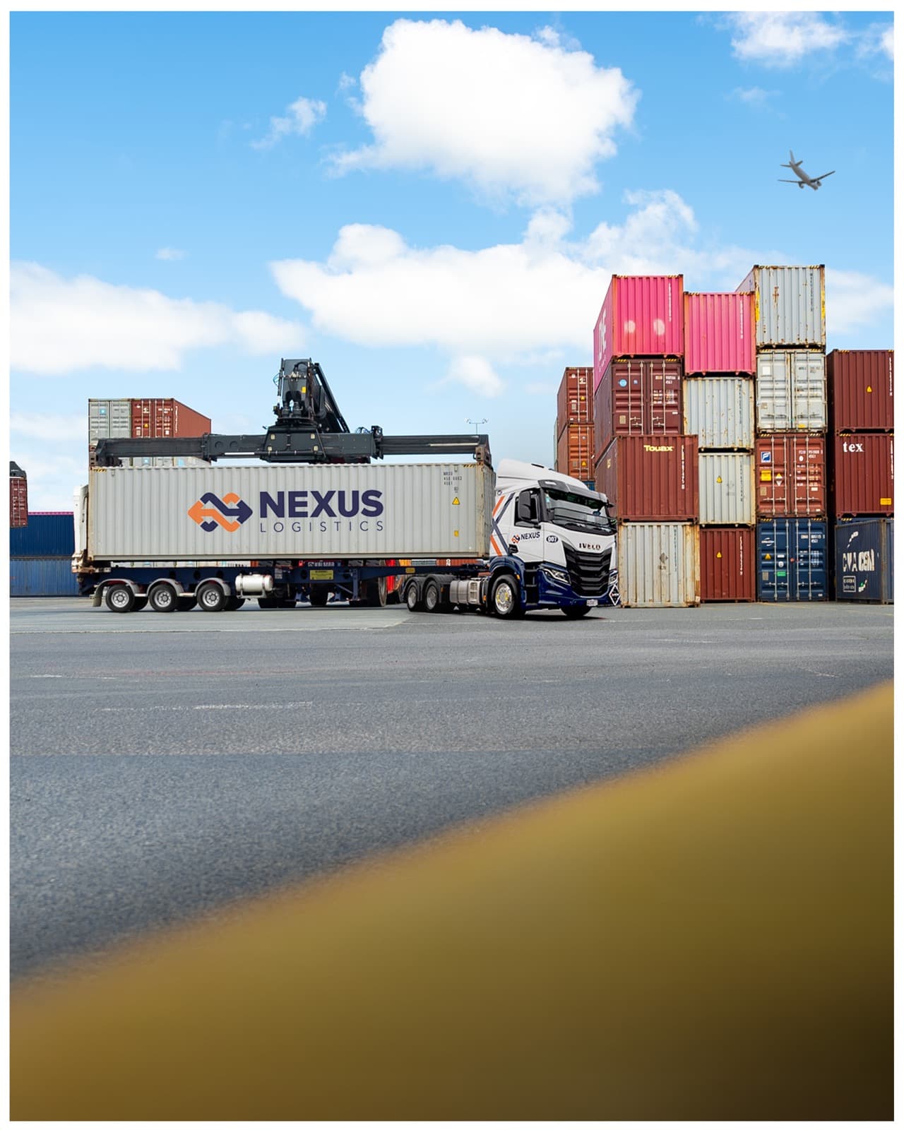 A Nexus Logistics branded truck parked in the container yard, with a plane overhead