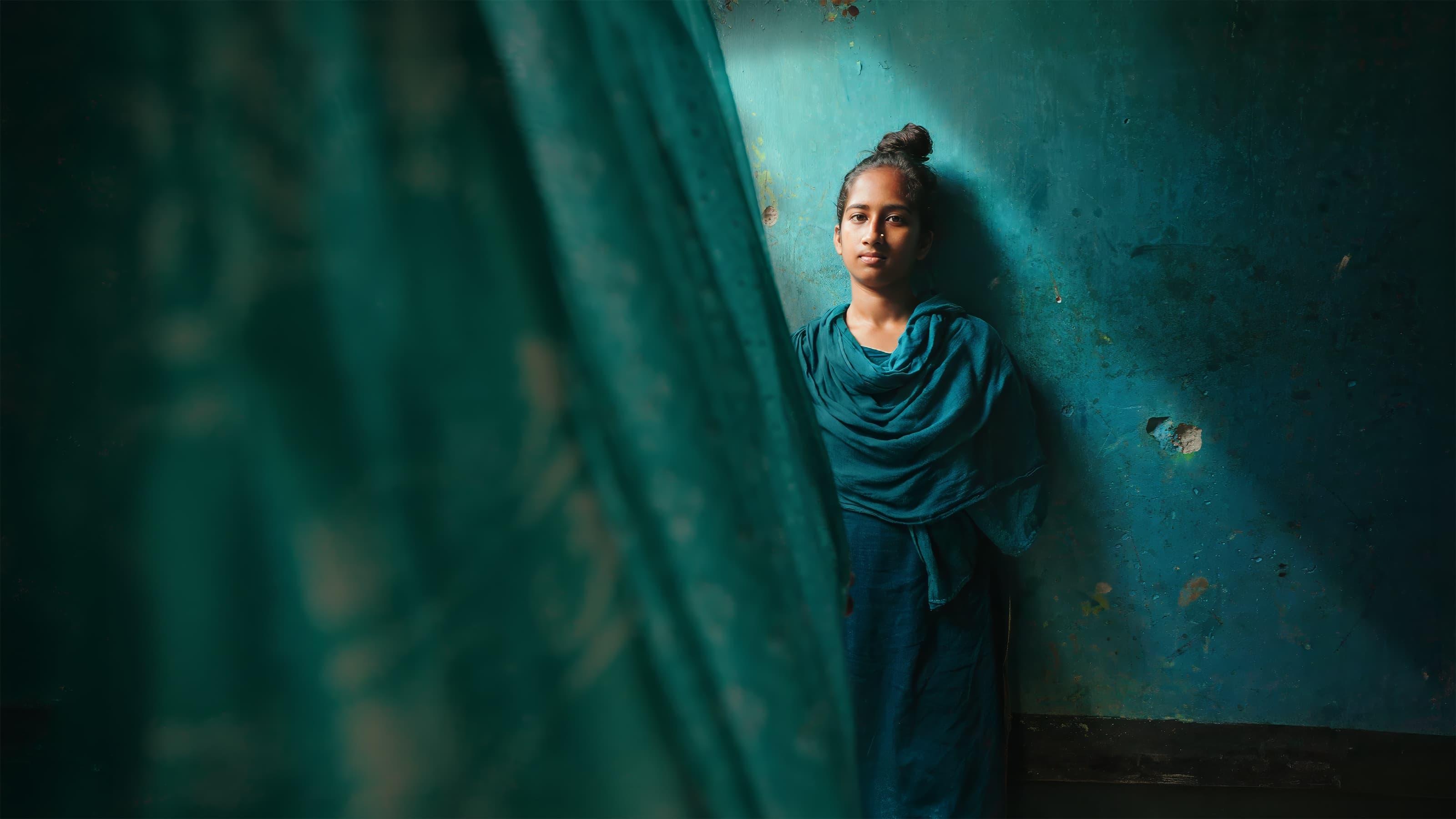 Portrait of a young woman in a teal shawl against a teal wall, partially framed by a curtain, Bangladesh