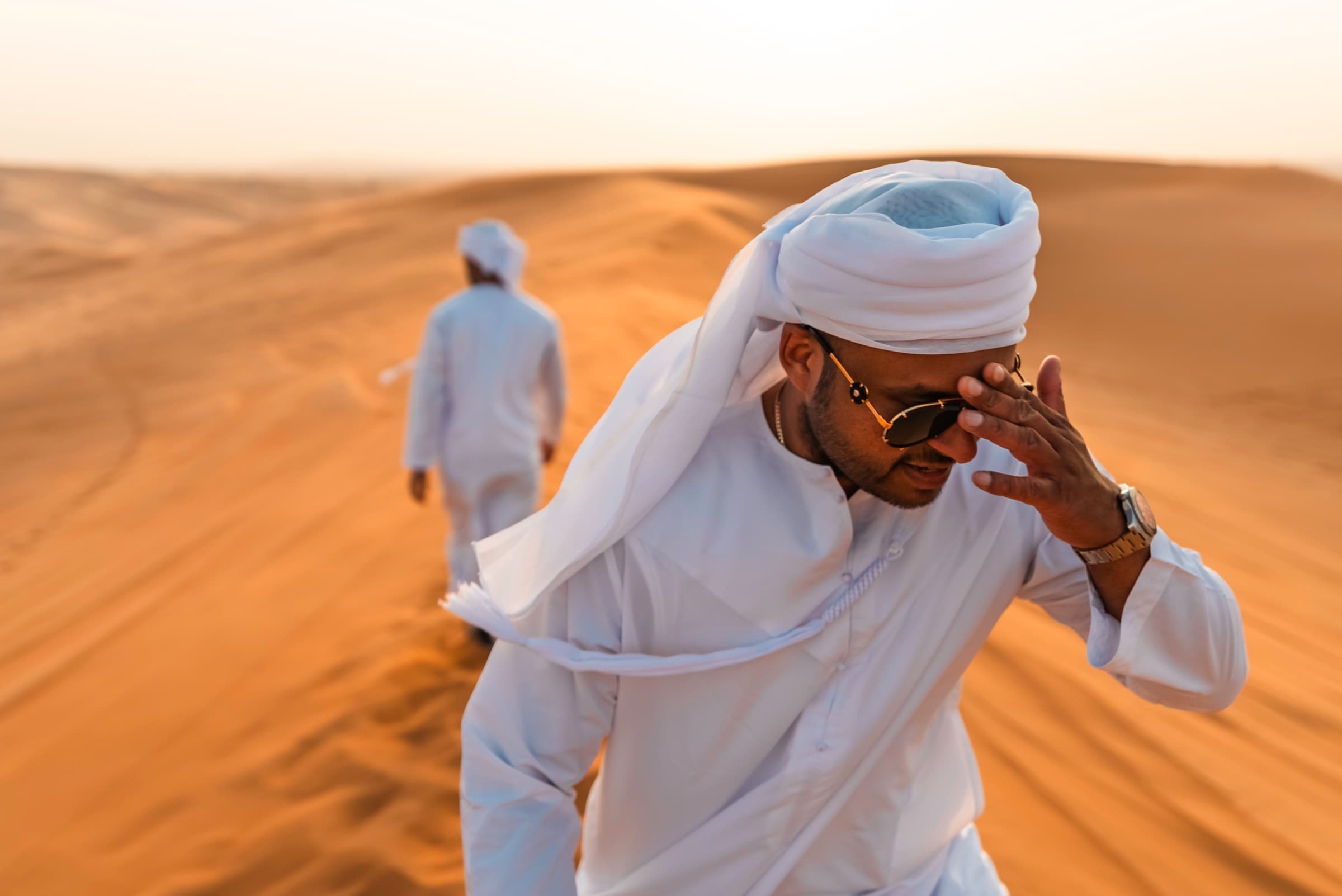 A man in traditional white Emirati dress shielding his eyes while walking across desert dunes