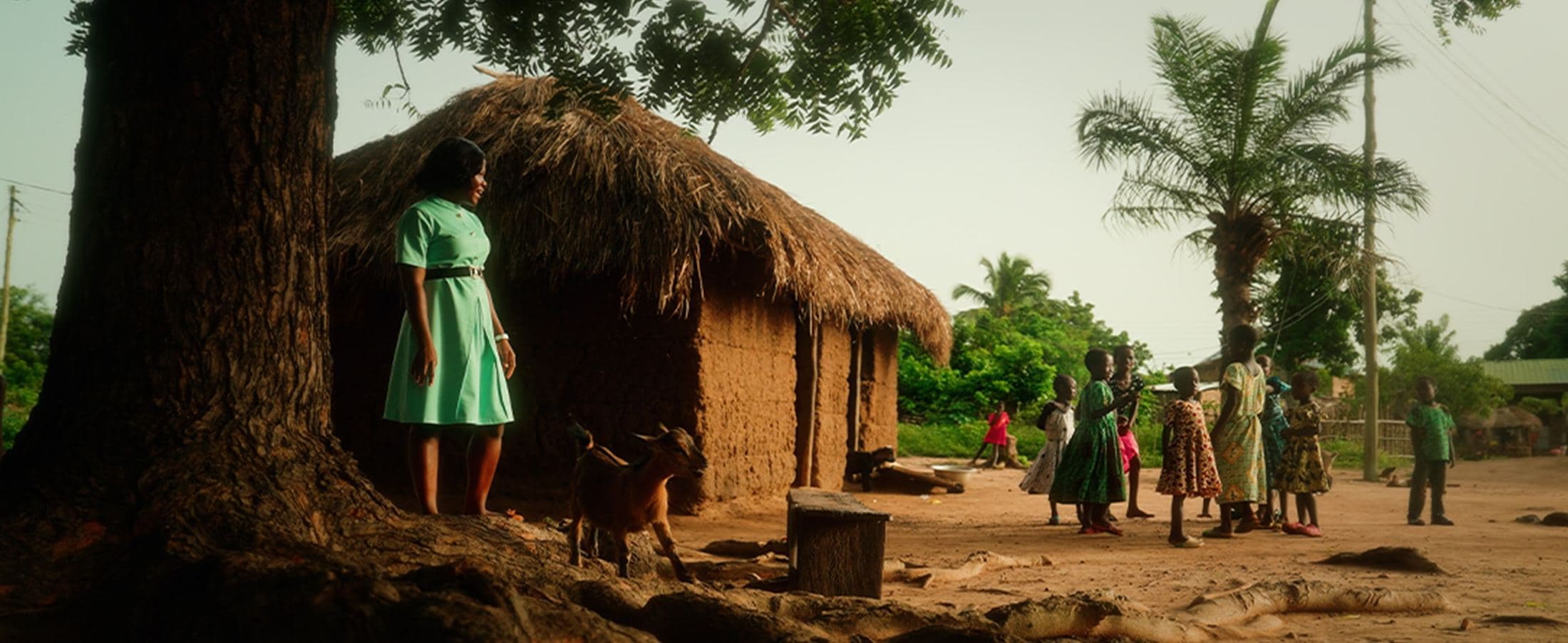 Girl standing beneath a tree by thatched huts, Ghana