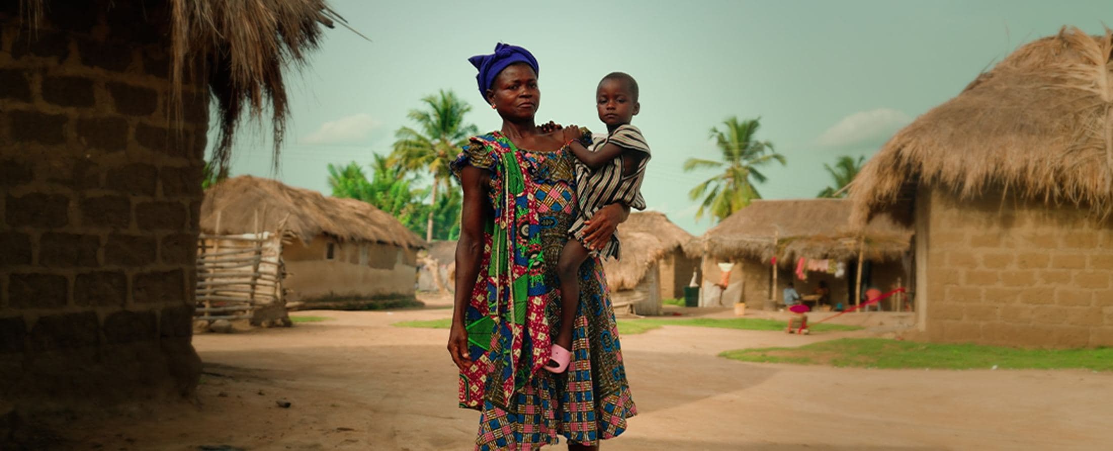 Mother carrying a child past thatched huts, Ghana